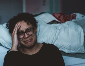 Erschöpfte Person mit Brille sitzt vor einem ungemachten Bett und hält sich den Kopf